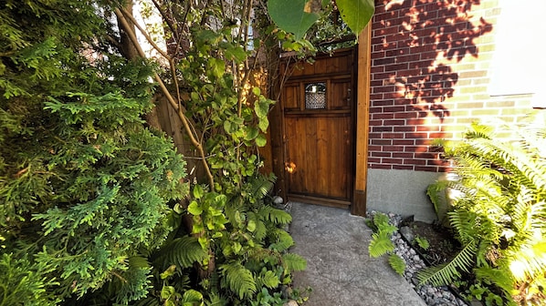 A wooden door with a small window, surrounded by dense greenery and a stone pathway leading to it.