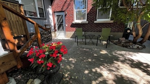 A wooden staircase leading to a door, a small outdoor seating area with chairs, and a potted plant with red flowers.