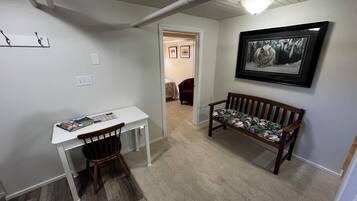 Un pasillo con un escritorio blanco, un banco de madera, un cuadro enmarcado en la pared y una puerta que da a otra habitación.