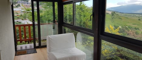 A balcony with a white chair and a wooden table, overlooking a green landscape with buildings in the distance.
