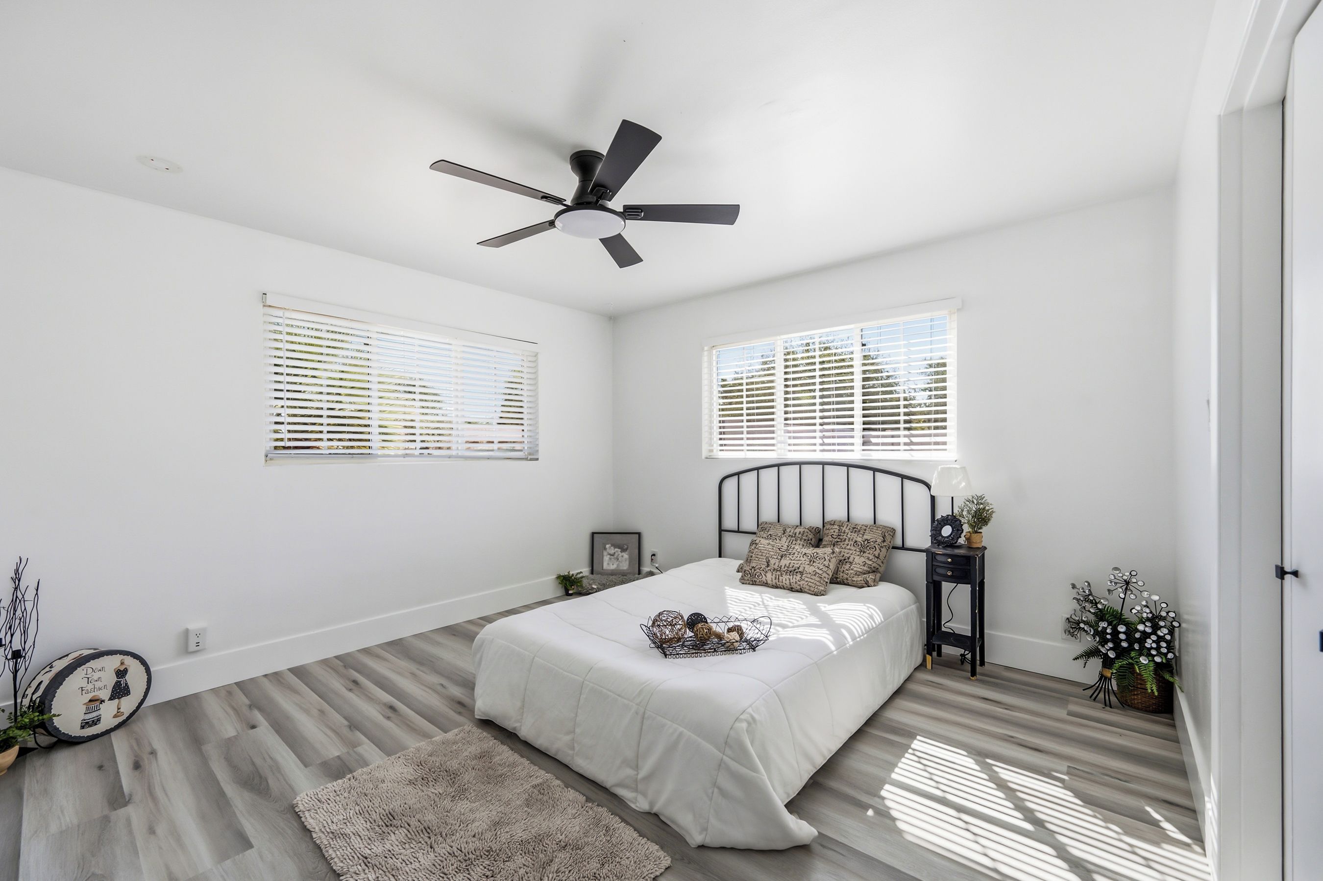 A bedroom with a bed, a ceiling fan, two windows with blinds, a bedside table with a lamp, and a small plant.