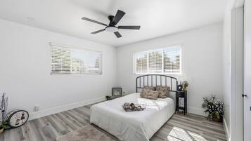 A bedroom with a bed, a ceiling fan, two windows with blinds, a bedside table with a lamp, and a small plant.
