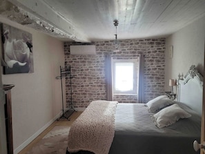 A bedroom with a stone wall, a bed with white bedding, a window with a view, and a ceiling fan. - Aponem Guesthouse, a charming 4-star property (Savignac-les-Églises)