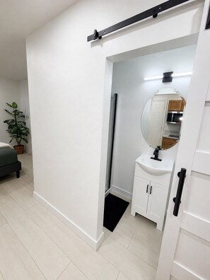 Ein modernes Badezimmer mit einem weißen Waschbecken, Spiegel und Schränken. Im angrenzenden Zimmer befindet sich ein Topf mit einer Pflanze und ein Bett.