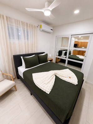 Ein Schlafzimmer mit Bett, Stuhl, Deckenventilator, Klimaanlage und einer Schiebetür zu einer Küchenzeile.