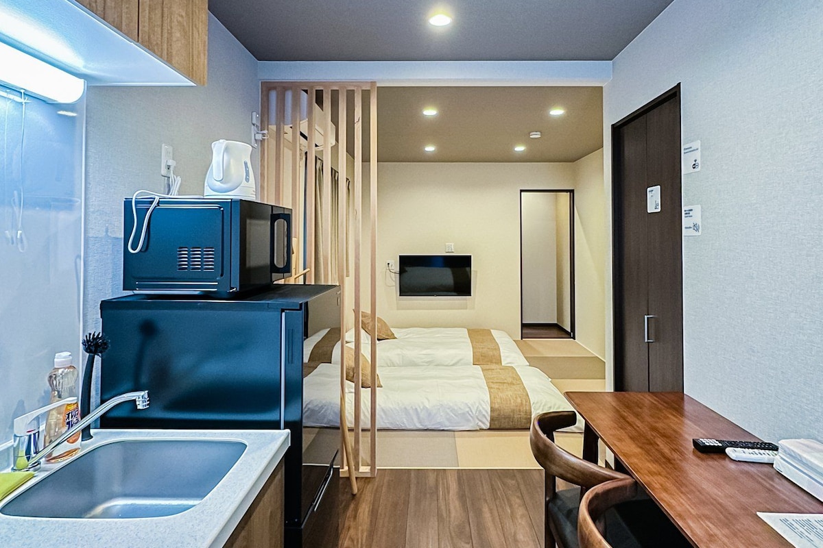 Ein kleines Hotelzimmer mit Küchenzeile, einem Bett, einem Fernseher und einem Holztisch.