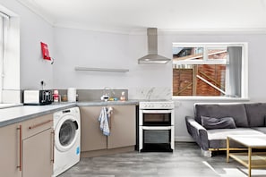 A compact kitchen with a washing machine, a stove, and a dining area with a sofa and table. - Old Trafford Studio – Walk to Tram, 10 Mins to Manchester Centre (Manchester)