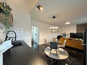 A modern kitchen with a black countertop, a dining area with a round table and chairs, and a living area with a sofa and a TV.