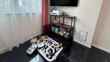 Ett vardagsrum med en platt-TV som är monterad på väggen, en svart möbel för underhållningsutrustning med diverse föremål, en mönstrad matta och ett fönster med tunna, genomskinliga gardiner.