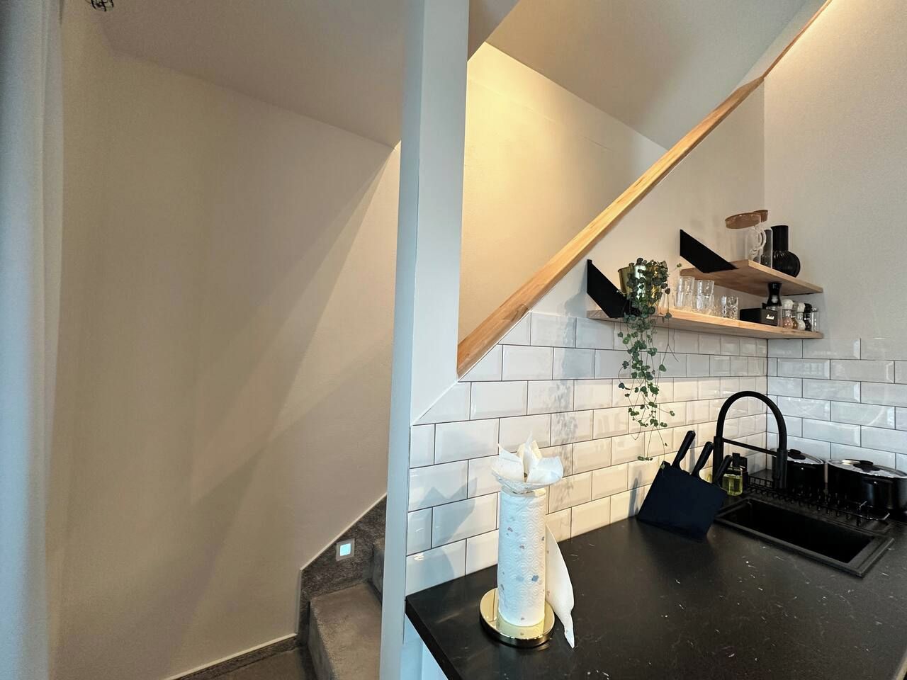 A kitchen with white tiled backsplash, a black countertop, and a wooden shelf with kitchen items.