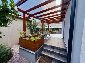 A covered outdoor area with a wooden bench, a table with a colorful tablecloth, and a garden bed with plants.