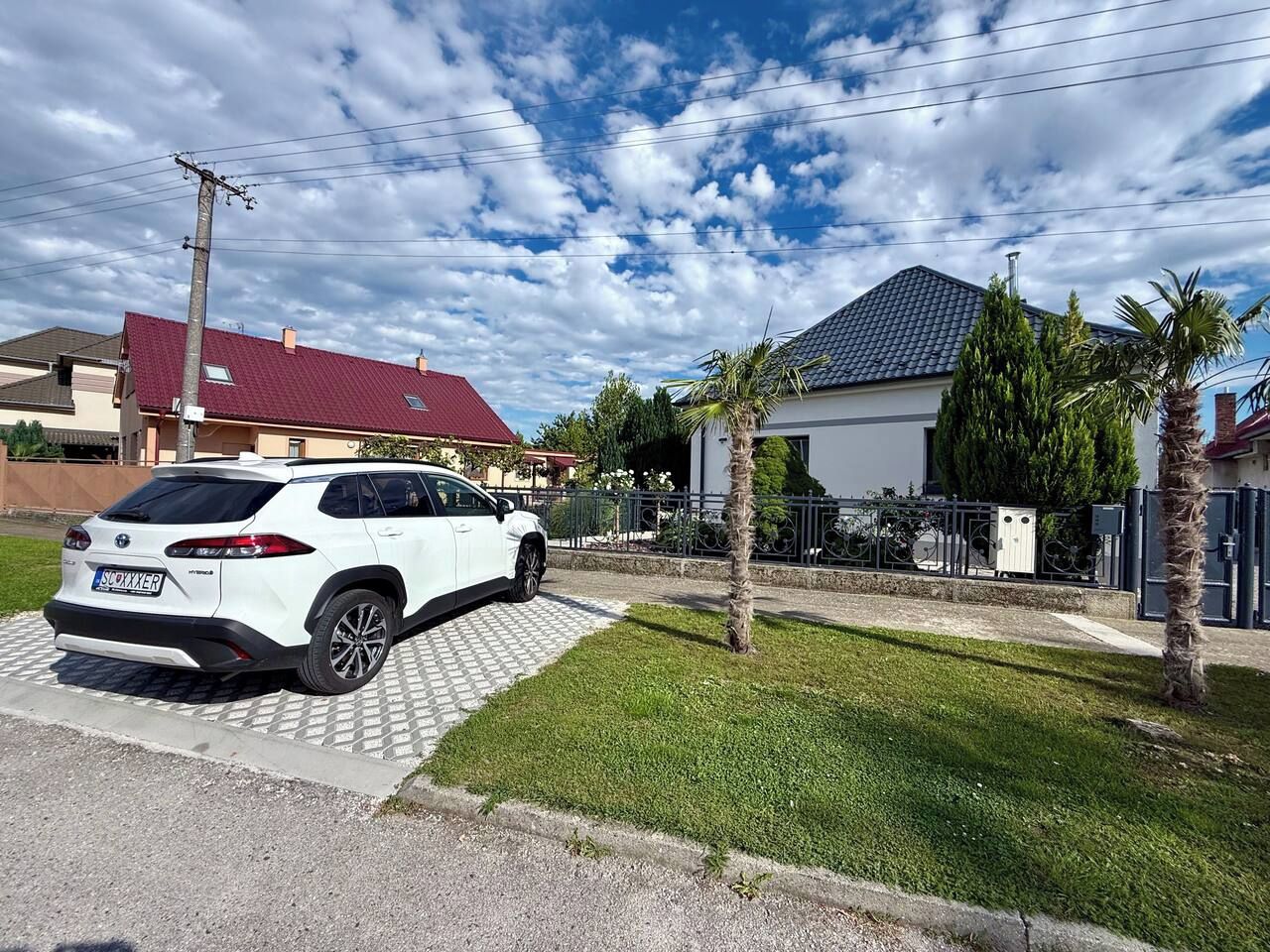 A white SUV parked on a paved driveway in a residential area with houses, a palm tree, and a clear sky.