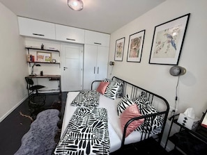 Un dormitorio con cama, mesita de noche y un escritorio con silla. Se observa un armario y una estantería con objetos encima.