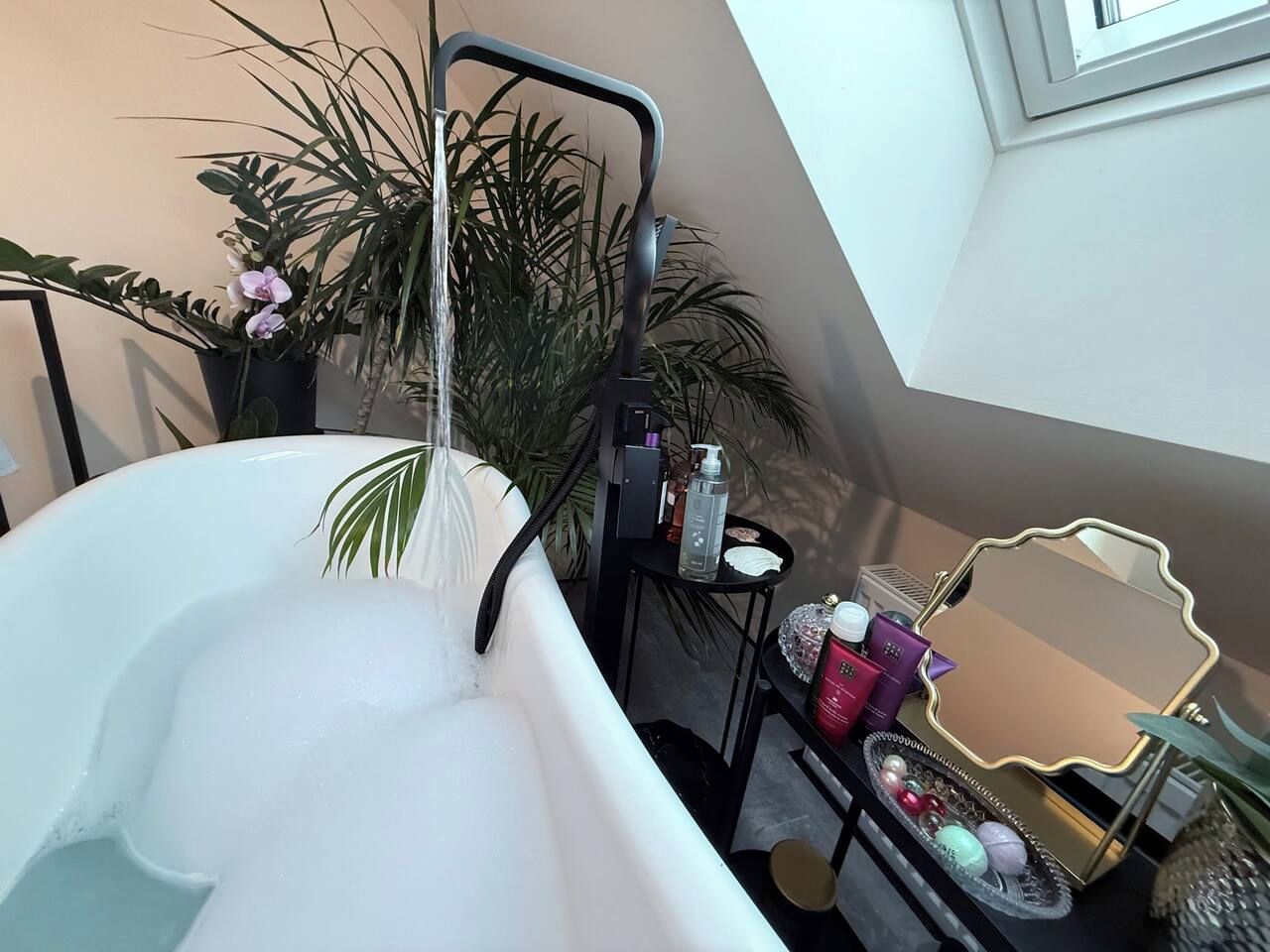 Een badkamer met een vrijstaand bad, een metalen plank in zwart met toiletartikelen, en een grote spiegel met gouden lijst.
