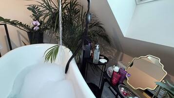 Een badkamer met een vrijstaand bad, een metalen plank in zwart met toiletartikelen, en een grote spiegel met gouden lijst.