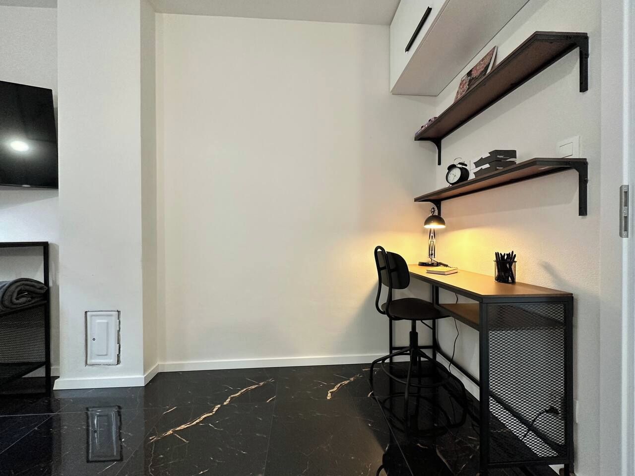Ein kleines Zimmer mit einem schwarzen Metallschreibtisch, einem Stuhl, einem kleinen Tisch mit einer Lampe und einem an der Wand befestigten Fernseher.
