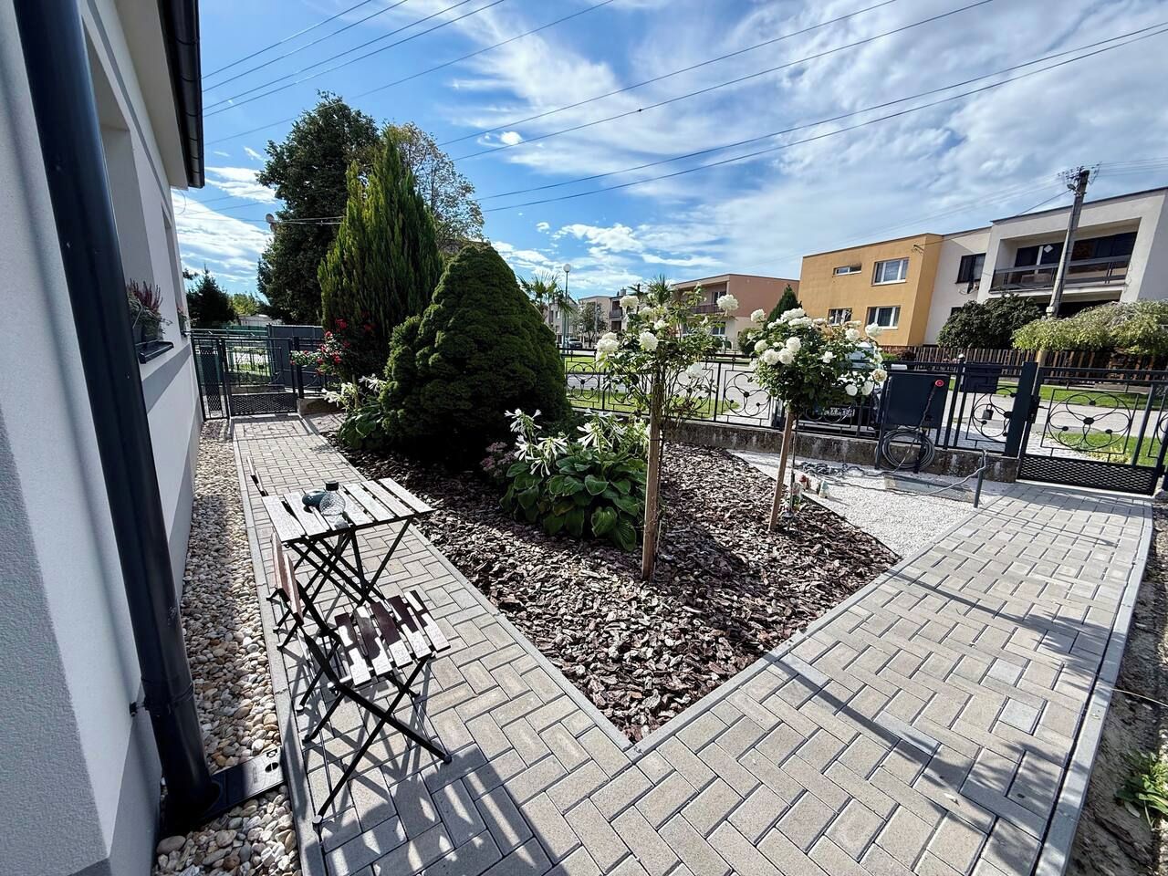 A paved outdoor area with a small garden bed, a metal bench, and a pathway leading to a building entrance.
