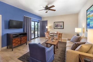 A hotel room with a flat-screen TV, a ceiling fan, a dining area with a table and chairs, a sofa, and a large window with curtains. - Royal Sea Cliff 2BR Resort Suite Kona Coast (Kailua-Kona)