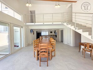 Ein geräumiger Essbereich mit einem langen Holztisch und Stühlen, einer Treppe mit Metallgeländer sowie Blick auf eine Küche und ein Wohnbereich.