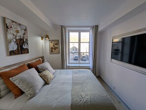 Quarto com cama, almofadas, televisão fixada na parede e janela com vista para carros estacionados.