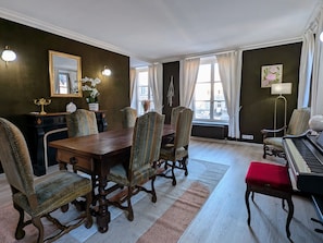 Uma sala de jantar com mesa e cadeiras de madeira, um piano e vista para a rua através de grandes janelas.