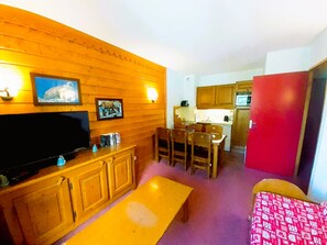 A room with a wooden wall, a television, a dining table with chairs, a kitchen area, and a red door. - Apartment with park view – 6 guests (Valfréjus)