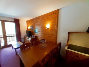 Taula de menjador de fusta amb cadires, una televisió i llums penjades a la paret en una habitació amb vistes a un balcó.