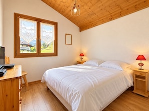 A bedroom with a wooden ceiling, a bed with white bedding, two bedside lamps, a desk with a computer, and a window with a view of a house and greenery. - Apartment facing Mont Charvin (Notre-Dame-de-Bellecombe)