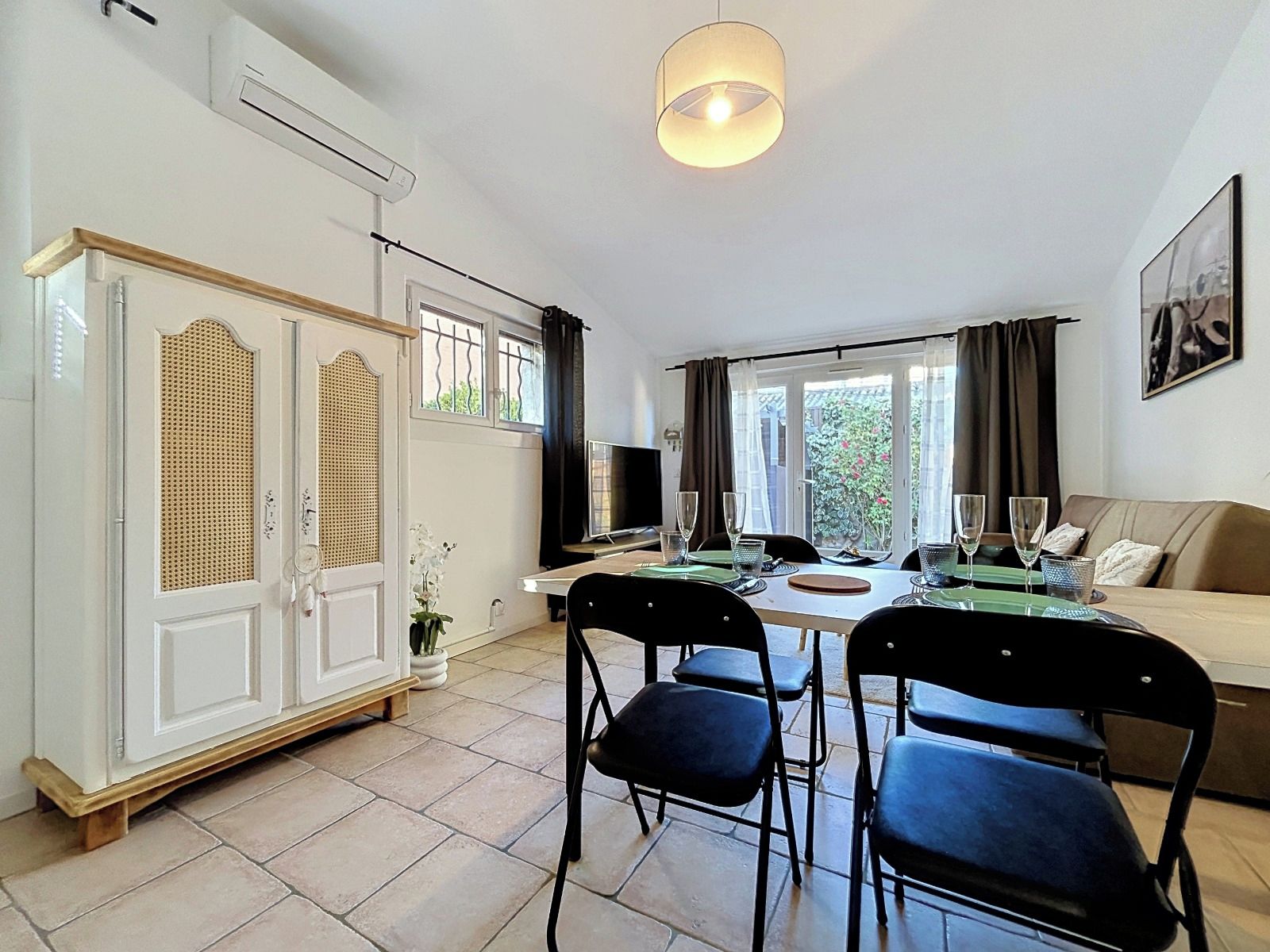 A well-lit room with a dining table, chairs, a sofa, a television, and a window with curtains.