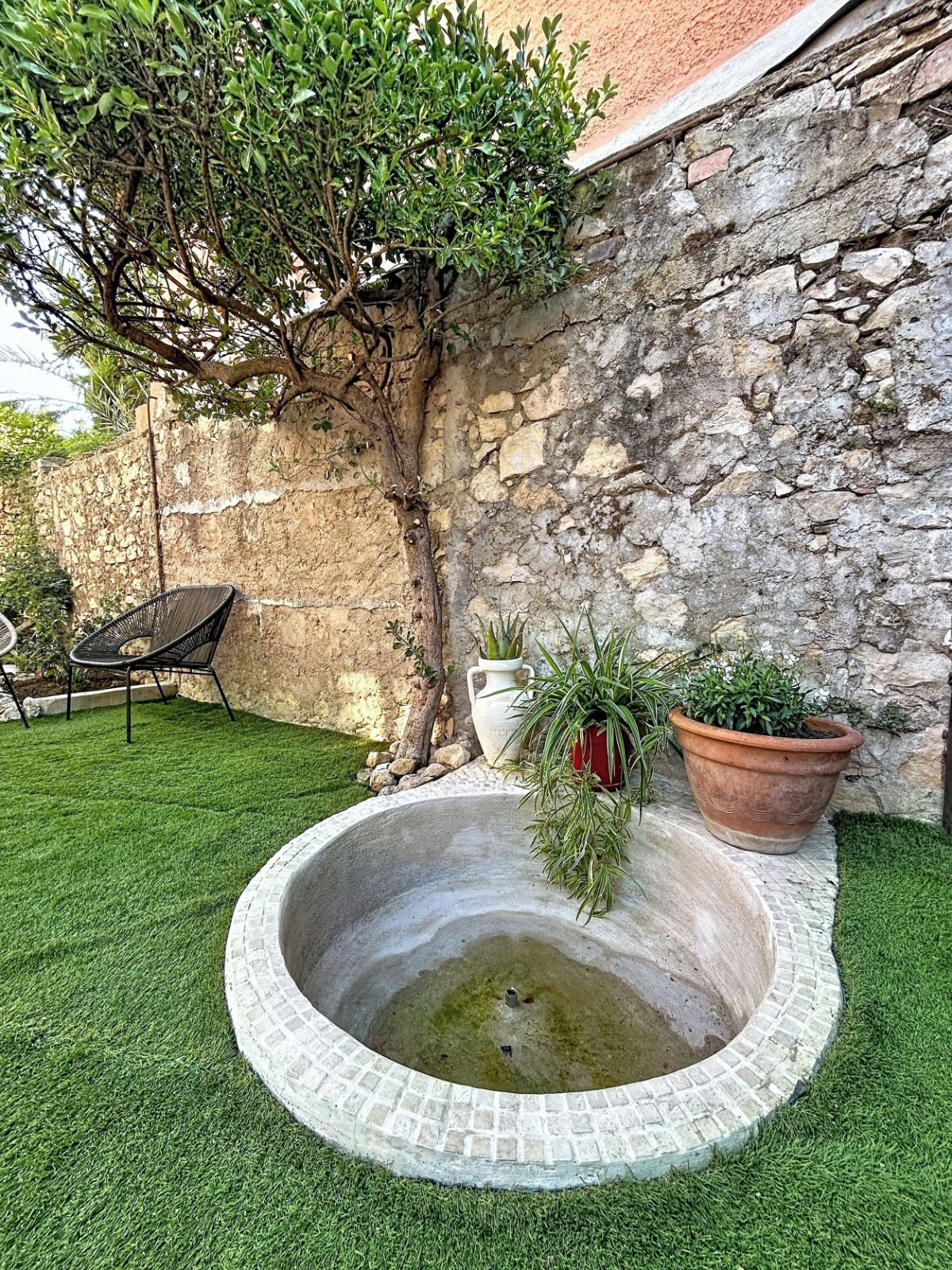 Un mur de pierre, une petite fontaine circulaire et des plantes en pot dans un espace aménagé en jardin.