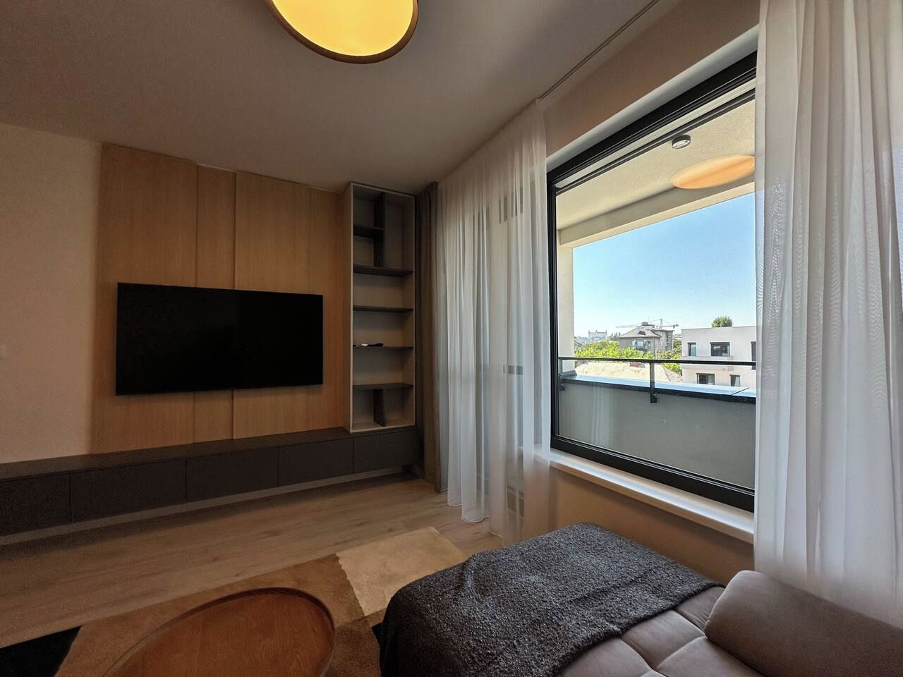 A modern hotel room with a flat-screen TV mounted on the wall, a sofa, a small table, and a window with a view of buildings.