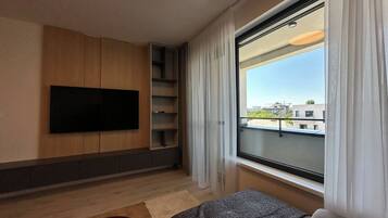 A modern hotel room with a flat-screen TV mounted on the wall, a sofa, a small table, and a window with a view of buildings.