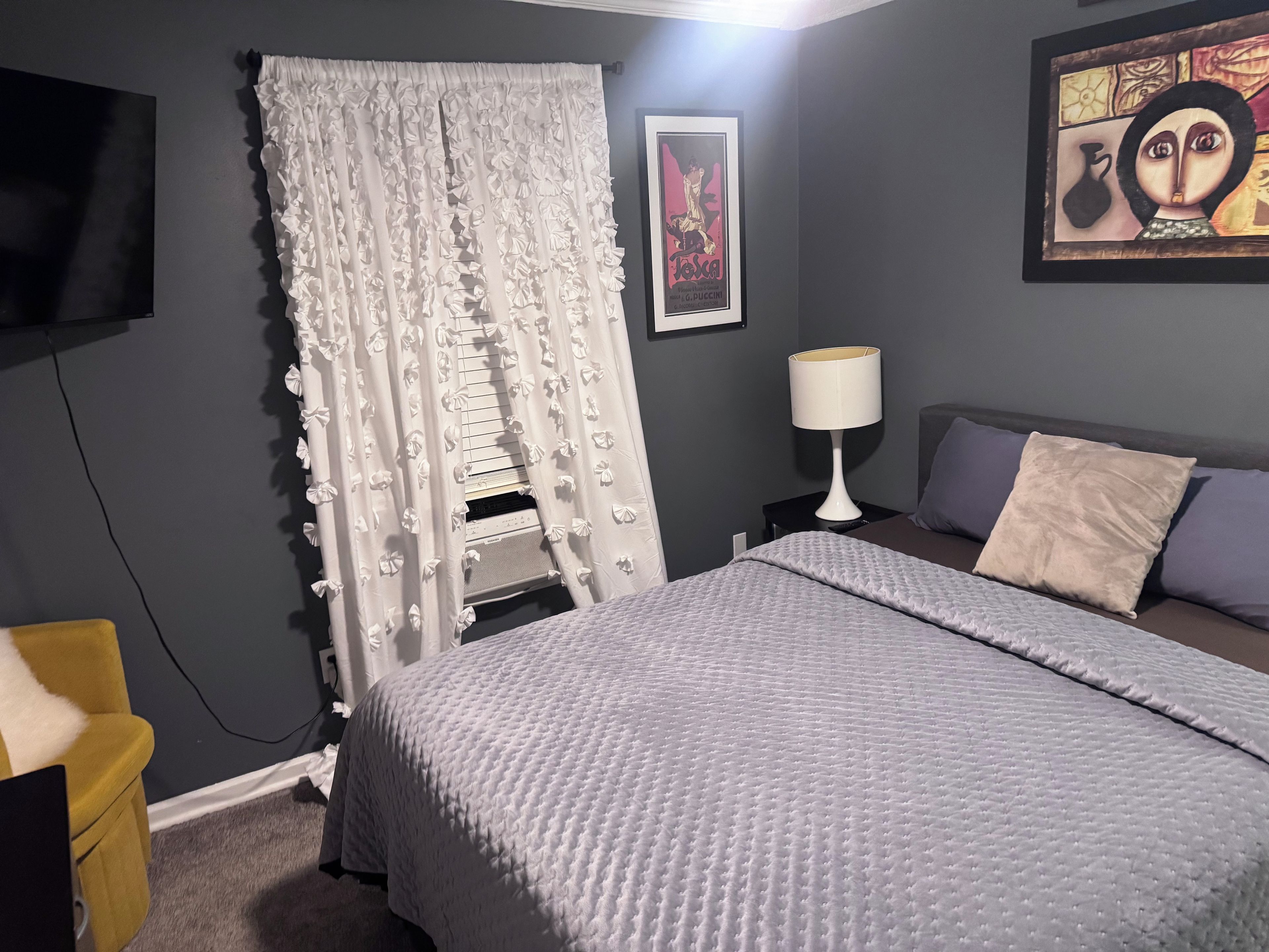 Een slaapkamer met een bed, een nachtkastje met een lamp, een aan de muur bevestigde televisie en een ingelijst schilderij.