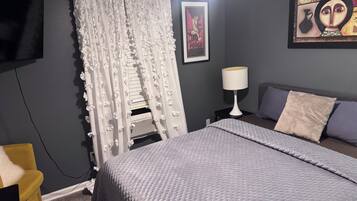 Een slaapkamer met een bed, een nachtkastje met een lamp, een aan de muur bevestigde televisie en een ingelijst schilderij.