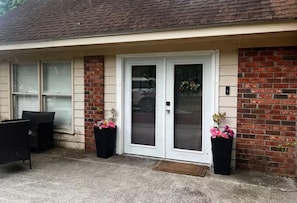 Un immeuble résidentiel avec une porte d’entrée, deux plantes en pot fleuries, un banc et un mur en briques.
