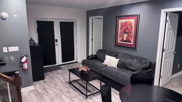Een moderne woonkamer met een grijze bank, een salontafel en een eettafel. Er zijn twee deuren, een brandblusser en een inlijsting met een afbeelding aan de muur.