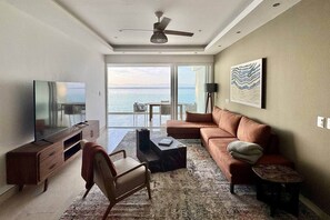 A modern living room with a large flat-screen TV, a sofa, a coffee table, and a chair. There is a balcony with a view of the sea. - Beachfront Luxury Condo - Steps from the Ocean (Bucerías)