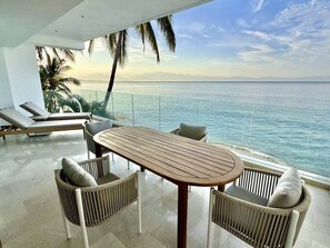 Ein Balkon mit Holztisch und Korbstühlen, der auf ein Gewässer mit Palmen und einem klaren Himmel blickt.