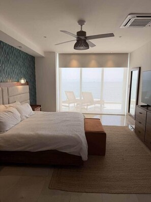 A modern bedroom with a large bed, a ceiling fan, a TV, and a view of the ocean. - Beachfront Luxury Condo - Steps from the Ocean (Bucerías)