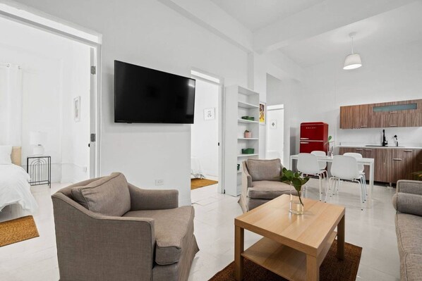 Un soggiorno moderno con una TV a schermo piatto, un divano, un tavolino da caffè e un'area pranzo con sedie, con una cucina sullo sfondo.