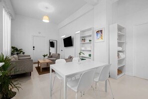 Un soggiorno moderno con un divano bianco, un tavolino da caffè e un tavolo da pranzo con sedie. C'è una TV montata a parete e una libreria con diversi oggetti.