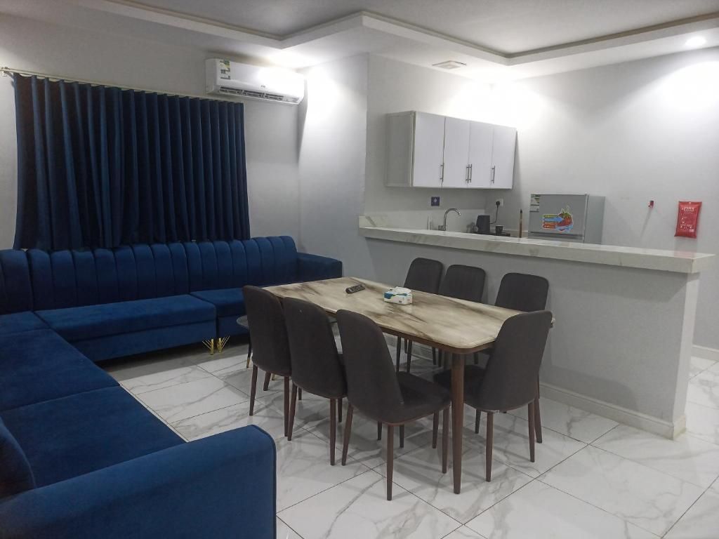 A modern living room with a blue sofa, a dining table with chairs, and a kitchen area with cabinets.