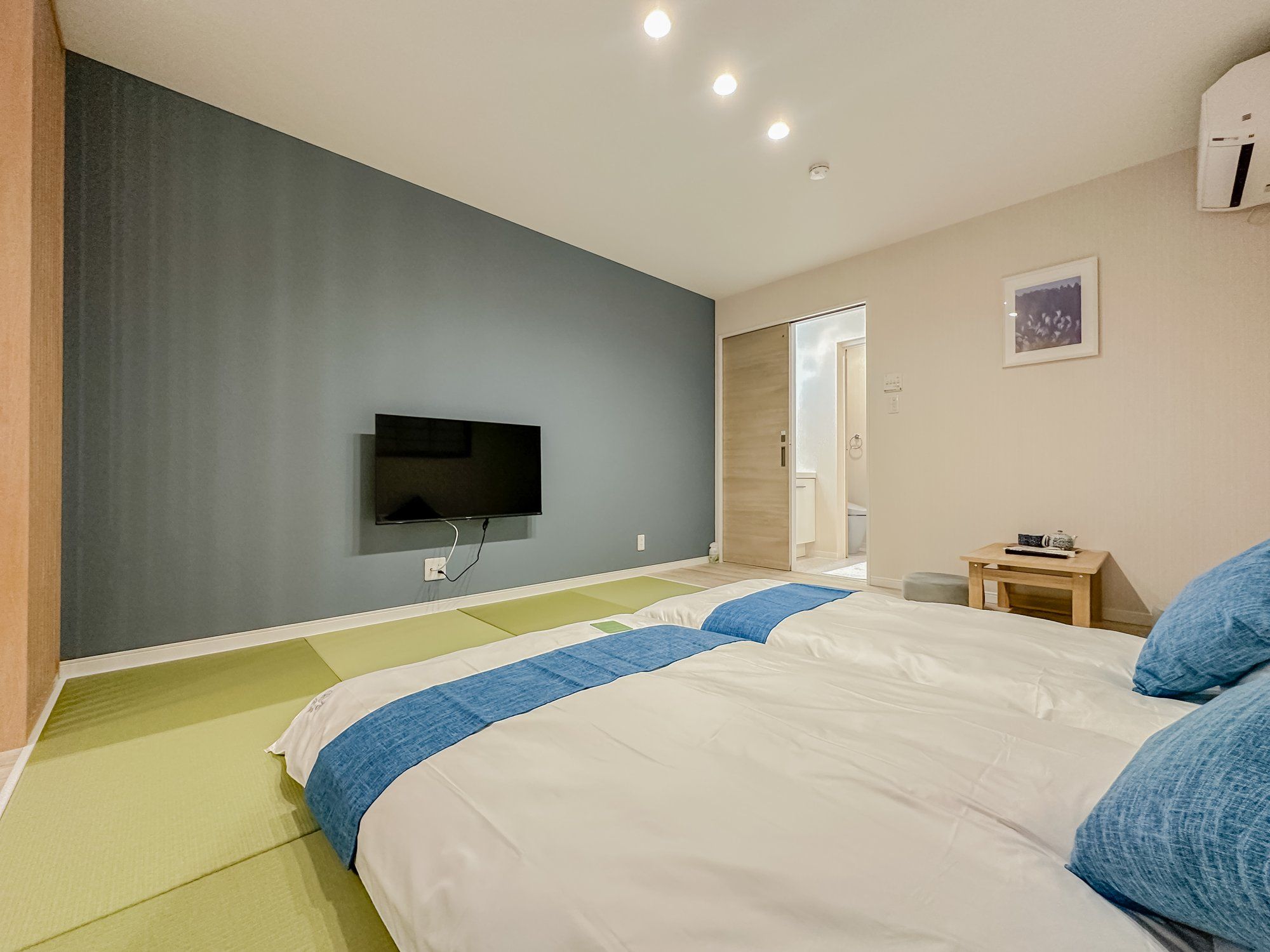 A hotel room with a bed, a television mounted on the wall, a small table with a cup, and a picture on the wall.