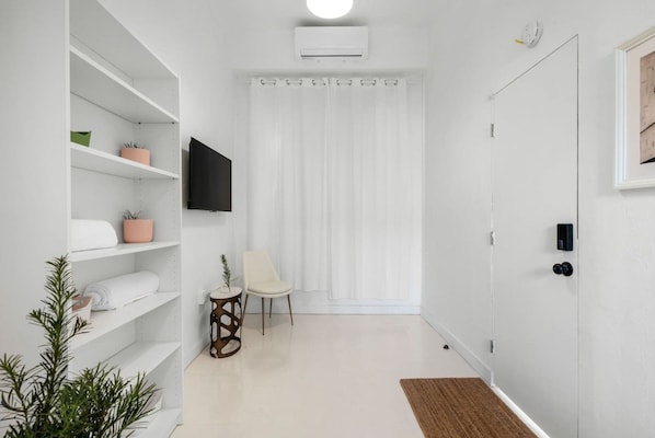 A modern, minimalistic room with white walls, a flat-screen TV mounted above a white shelf, a white chair, and a door with a handle. - Casa San Rafael Apt 2 | Aesthetic Escape for Two (San Juan de Puerto Rico)