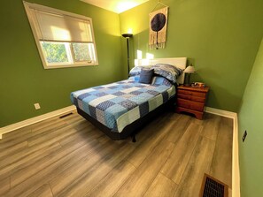 A bedroom with a bed, a nightstand, a lamp, a window, and a wall decoration. - Cheerful 3 bedroom waterfront cottage with WiFi Tv (Selwyn)