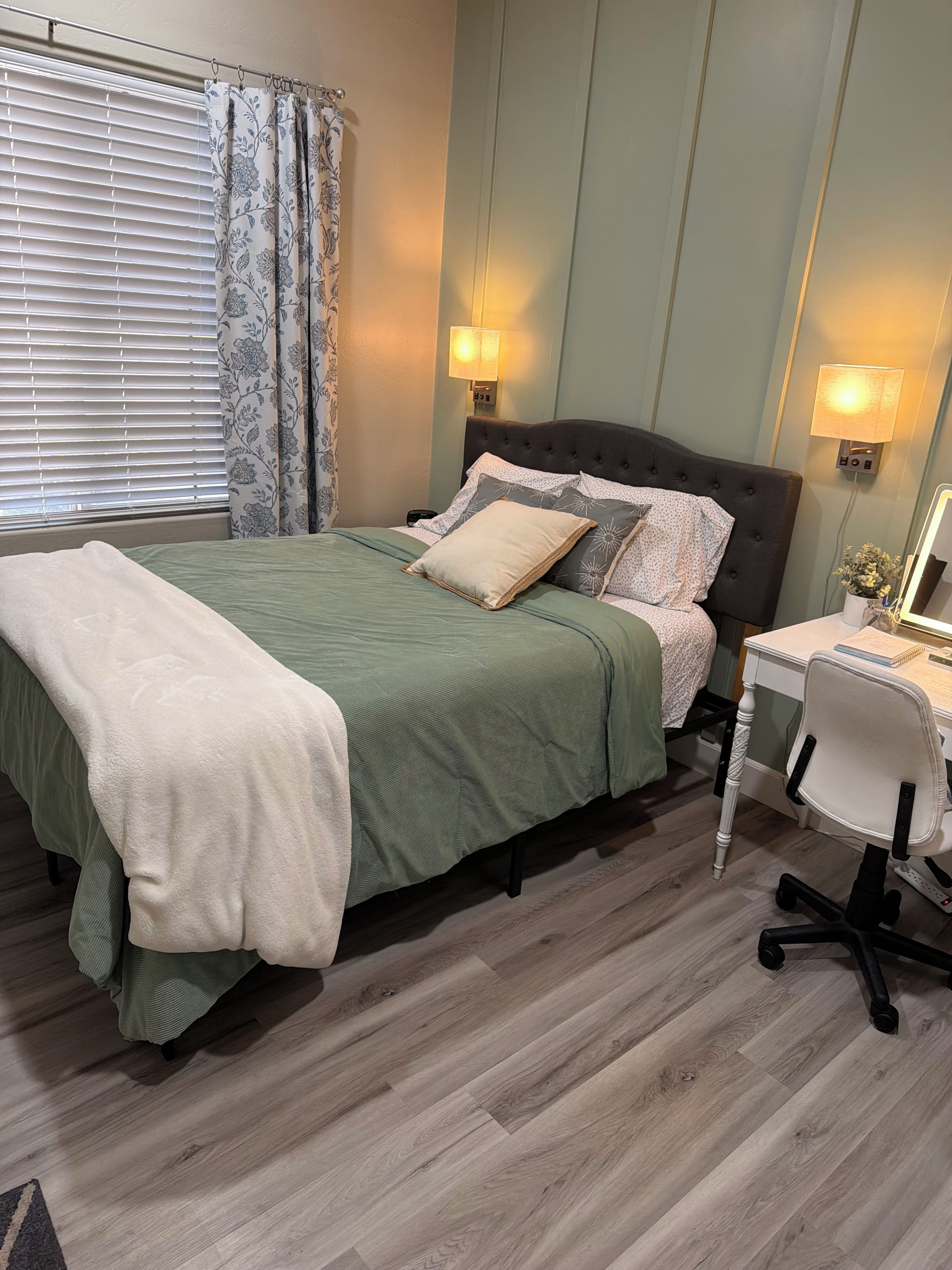 A bedroom with a bed, bedside lamps, a desk with a chair, and a window with blinds and curtains.