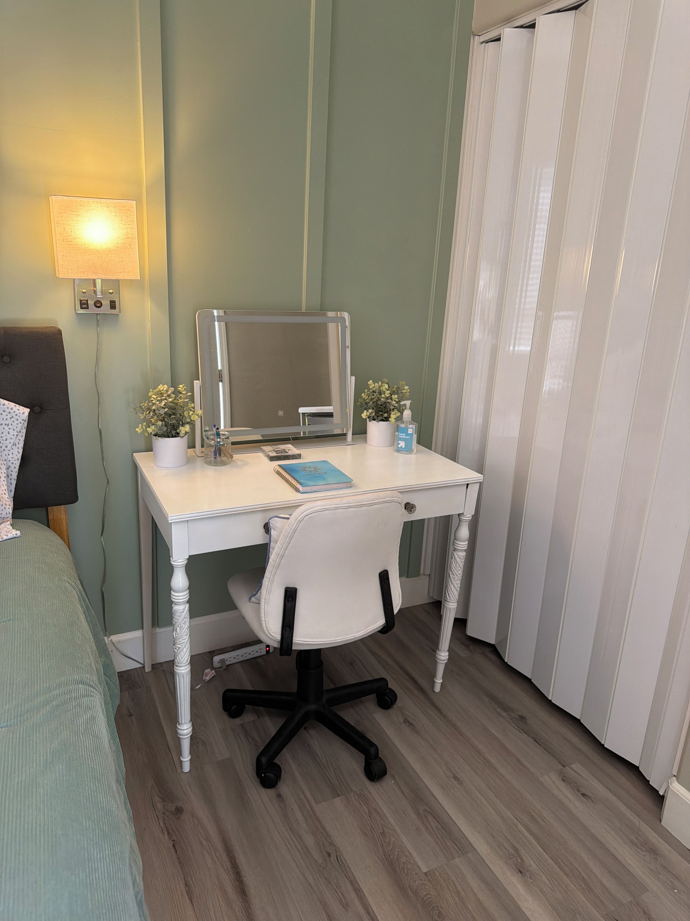Una habitación con un escritorio blanco, una silla, una cama, una lámpara, un espejo y una planta en maceta.