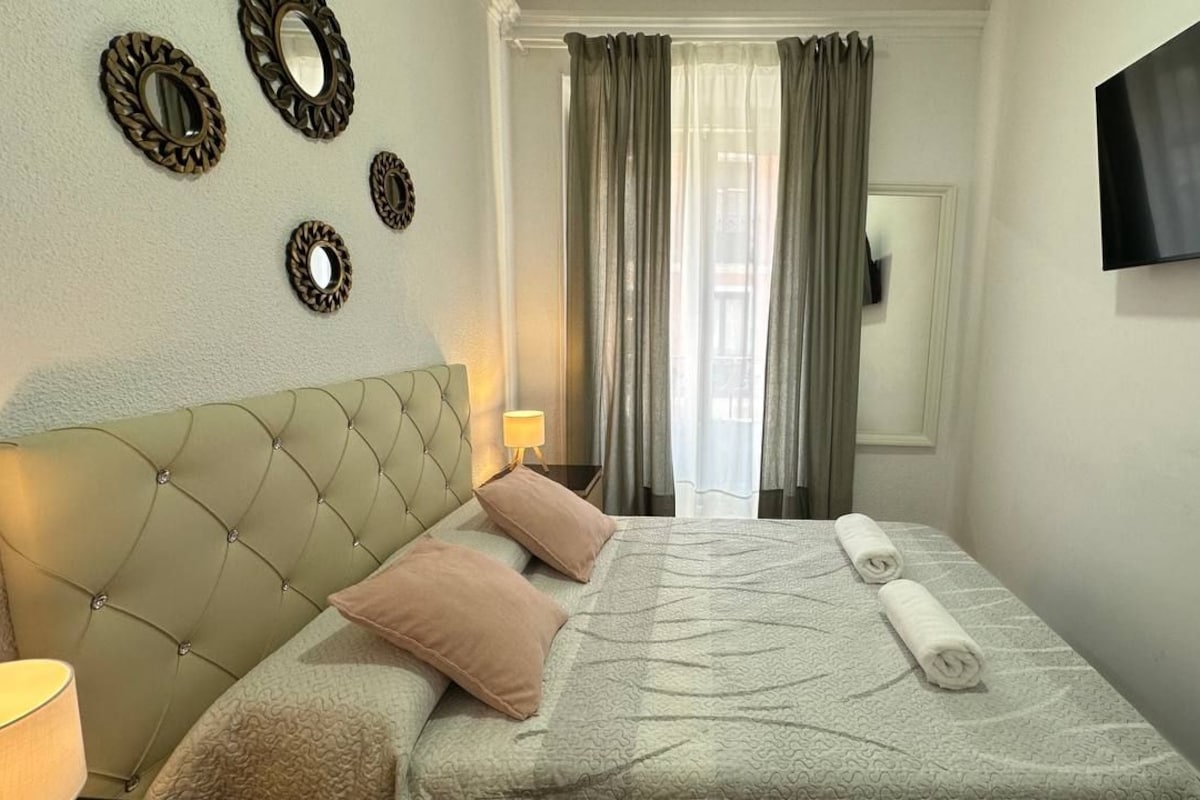 A hotel room with a tufted headboard, a bed with a patterned bedspread, two pillows, a rolled towel, a bedside lamp, a flat-screen TV mounted on the wall, and a window with curtains.