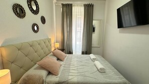 A hotel room with a tufted headboard, a bed with a patterned bedspread, two pillows, a rolled towel, a bedside lamp, a flat-screen TV mounted on the wall, and a window with curtains. - Habitaciones Malasaña (Madrid)