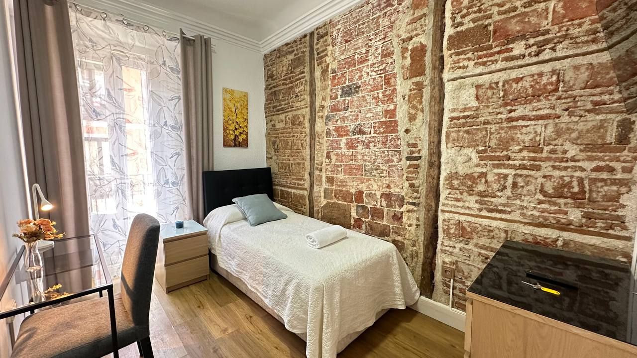 Un dormitorio con pared de ladrillo, cama con cubrecama blanco, escritorio con silla y ventana con cortinas.